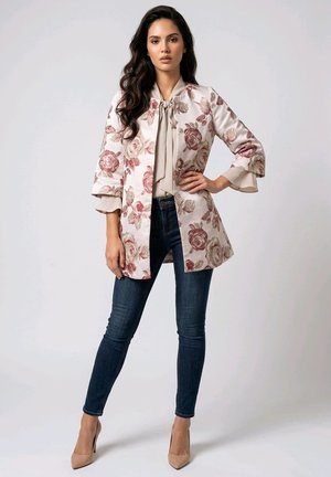 Mujer de pie con la mano en la cadera, vistiendo un abrigo con estampado floral, blusa beige con lazo, vaqueros ajustados oscuros y tacones altos de color nude.