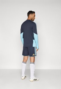 Camisola de manga longa azul-marinho com mangas azul claro, combinada com calções azul-marinho com detalhes em dourado e três riscas. Meias brancas e botas de futebol.