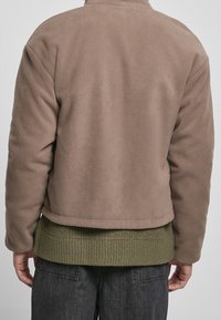 Giacca in pile marrone con colletto alto, design accorciato e orlo lavorato in maglia verde oliva, abbinata a pantaloni grigi scuri.