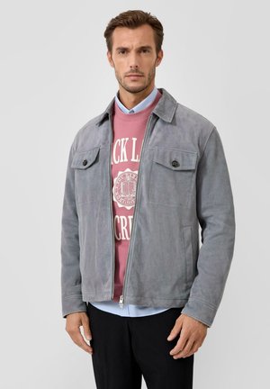 Mężczyzna w szarej zamszowej kurtce na różowej bluzie z nadrukiem oraz jasnoniebieskiej koszuli z kołnierzykiem, stojący na jednolitym tle.