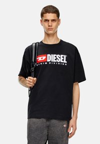 Melnas kokvilnas T-krekls ar īsām piedurknēm, ar balto un sarkano "DIESEL" logotipu un "DENIM DIVISION" tekstu, kas drukāts priekšpusē.