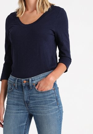 Vrouw met een donkerblauw shirt met lange mouwen en lichtblauwe spijkerbroek met één hand in haar zak tegen een witte achtergrond.