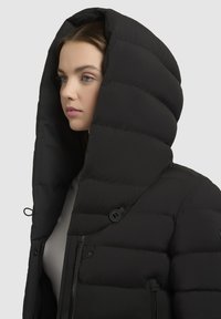 Vrouw in een zwart gewatteerd winterjack met een grote capuchon, die naar links kijkt tegen een lichtgrijze achtergrond.