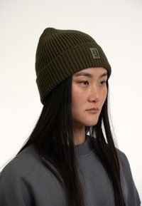 Caciula tricotată verde, cu nervuri, având un tiv pliat și un patch cu logo rectangular. Poartă o bluză gri, cu un guler înalt și mâneci lungi.