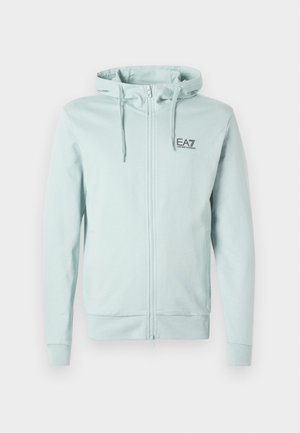 Haine à capuche zippée bleu clair en tissu doux, avec un capuchon à cordon, des poches latérales et un petit logo noir sur la poitrine.