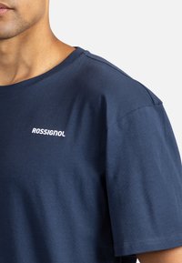 Tmavě modré bavlněné tričko s kulatým výstřihem, krátkými rukávy a bílým logem "ROSSIGNOL" na levé straně hrudi. Hladká textura, ležérní design.