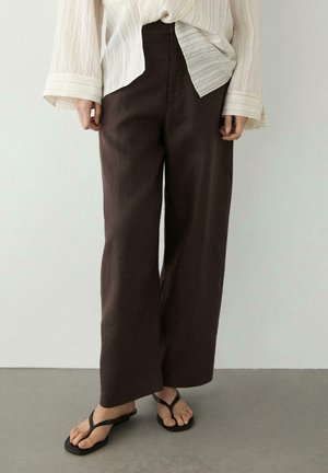 Persoon met losse donkerbruine broek, wit textuur lang mouwen blouse en zwarte teenslippers, staand op een grijze vloer.