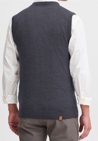 Pull en tricot sans manches gris foncé avec ourlet et col côtelés. Porté sur une chemise à manches longues blanche, mettant en valeur un motif en tricot texturé.