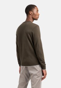 Maglione verde oliva con scollatura rotonda e orlo a costine. Texture liscia, maniche lunghe, indossato sopra pantaloni beige chiaro.