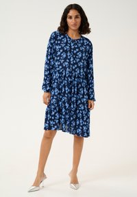 Robe bleu marine avec un motif floral bleu clair, manches longues, bouton sur le devant, jupe à étages, fabriquée en tissu léger, associée à des talons argentés.
