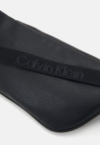 Musta nahkalaukku, jossa on teksturoitu pinta ja voimakkaasti esiin tuleva Calvin Klein -logo, punottu hihna edessä. Sujuva, hieman kaareva muoto.