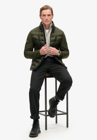 Ciemnozielona ocieplana kurtka z wysokim kołnierzem i dwiema zasuwanymi kieszeniami, nałożona na jasnoszary sweter. Czarne spodnie i buty dopełniają stylizację.