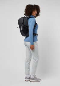 Schwarzer Rucksack mit einem schlanken Design und verstellbaren Trägern, getragen über einem blauen Langarmshirt und hellgrauen Hosen, kombiniert mit grauem Schuhwerk.