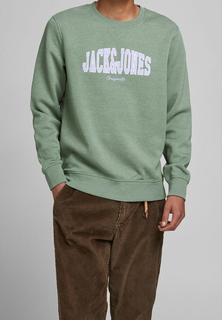 Personne portant un sweat vert sauge avec le texte "JACK&JONES Originals" et un pantalon en velours côtelé marron, mains partiellement dans les poches, sur un fond gris.