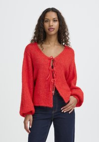 Maglione rosso in maglia pelosa con scollo a V, dettaglio con nodo frontale e maniche oversize. Indossato con jeans scuri, caratterizzati da una vestibilità comoda e un tessuto strutturato.