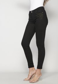 Black skinny jeans with intricate gold embroidery on front pockets, featuring a classic button and zip closure. Smooth texture, form-fitting design.