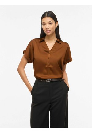 Moteris dėvi trumpomis rankovėmis rudos spalvos satininę palaidinę, įkištą į juodus aukšto liemens kelnes su juodu diržu, stovinti su rankomis kišenėse.