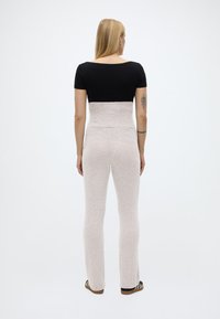 Γυναίκα με μακριά ξανθά μαλλιά, γυρισμένη πλάτη, φορεί μια μαύρη μπλούζα, μπεζ παντελόνι ψηλόμεσο και ριγέ επίπεδα παπούτσια, σε ουδέτερο λευκό φόντο.