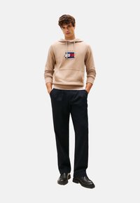 Joven vistiendo un hoodie beige con logo, pantalones negros y zapatos negros, de pie con las manos en los bolsillos frente a un fondo liso.