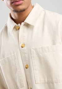 Crème kleurige, knoopsluiting shirt gemaakt van katoen. Beschikt over een klassieke kraag, twee voorzakken en gele knopen. Textuur oppervlak.