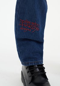 Pierna de pantalón de mezclilla azul oscuro con texto bordado en rojo "Diseños de Don Ed Hardy" junto a una bota negra con cordones.