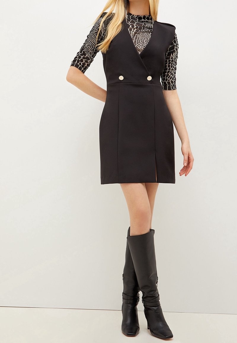 Femme portant une robe noire sans manches avec des boutons argentés sur un haut à manches longues à motifs, associée à des bottes noires jusqu'au genou, se tenant les jambes croisées.
