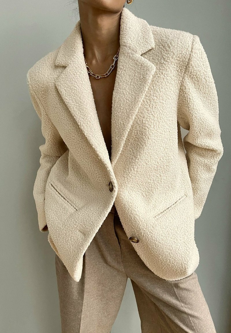 Blazer oversize de couleur crème avec tissu texturé, col à revers cranté, fermeture à un bouton et poches avant. Associé à un pantalon beige.