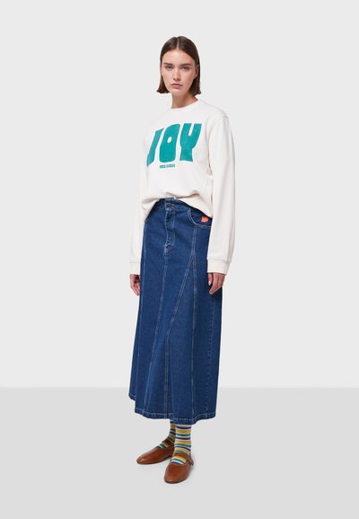 Felpa bianca con grafica teal, abbinata a una lunga gonna di jeans blu. Scarpe marroni e calze a righe completano l'outfit.