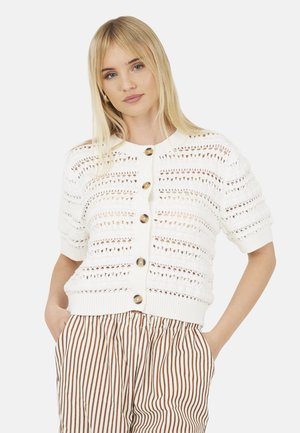 Jeune femme aux longs cheveux blonds portant un cardigan en maille blanc boutonné et un pantalon taille haute à rayures verticales marron et blanches.