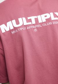 Camiseta de algodón marrón con texto blanco en negrita que dice "MULTIPLY APPAREL CLUB 2020" en la parte trasera, con un acabado liso y ligeramente texturizado.