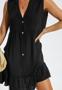 Robe noire sans manches en matériau texturé avec un col en V, quatre boutons et un ourlet à volants. Associée à un sac à main tissé.