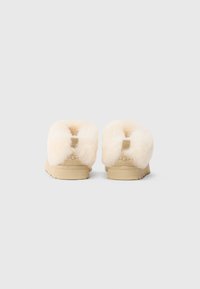 Une paire de pantoufles beige à enfiler avec une doublure en laine de mouton duveteuse vue de l'arrière sur un fond uni.