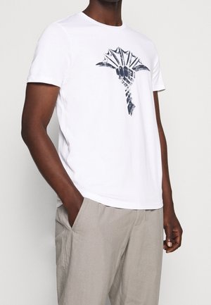 Weißes Baumwoll-T-Shirt mit dunklem Grafikdesign auf der Vorderseite, kurzen Ärmeln und rundem Ausschnitt, kombiniert mit hellgrauen Hosen.