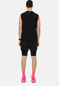 Top atlético negro sin mangas, pantalones cortos negros sobre mallas compresoras, calcetines rosas con zapatillas, visto desde atrás contra un fondo blanco.