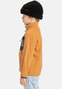 Hnědá fleecová bunda s vysokým límcem, černou přední kapsou a šedými akcenty, nošená s černou pletenou čepicí. Světle modré džíny dotvářejí celý outfit.