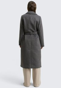 Vrouw in een lange, gebelde grijze jas over wijde beige broek, staande met haar gezicht naar weggekerd tegen een effen lichte achtergrond.