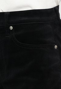 Κοντινό πλάνο τσέπης μαύρου βελούδινου παντελονιού με ασημένιες λεπτομέρειες και ραφές, φορεμέ�νο με λευκό πουκάμισο μέσα από το παντελόνι.