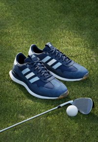Scarpe da golf blu navy con tomaia in suede e materiale sintetico, tre strisce bianche e intersuola bianca. Include una mazza da golf e una pallina in primo piano.