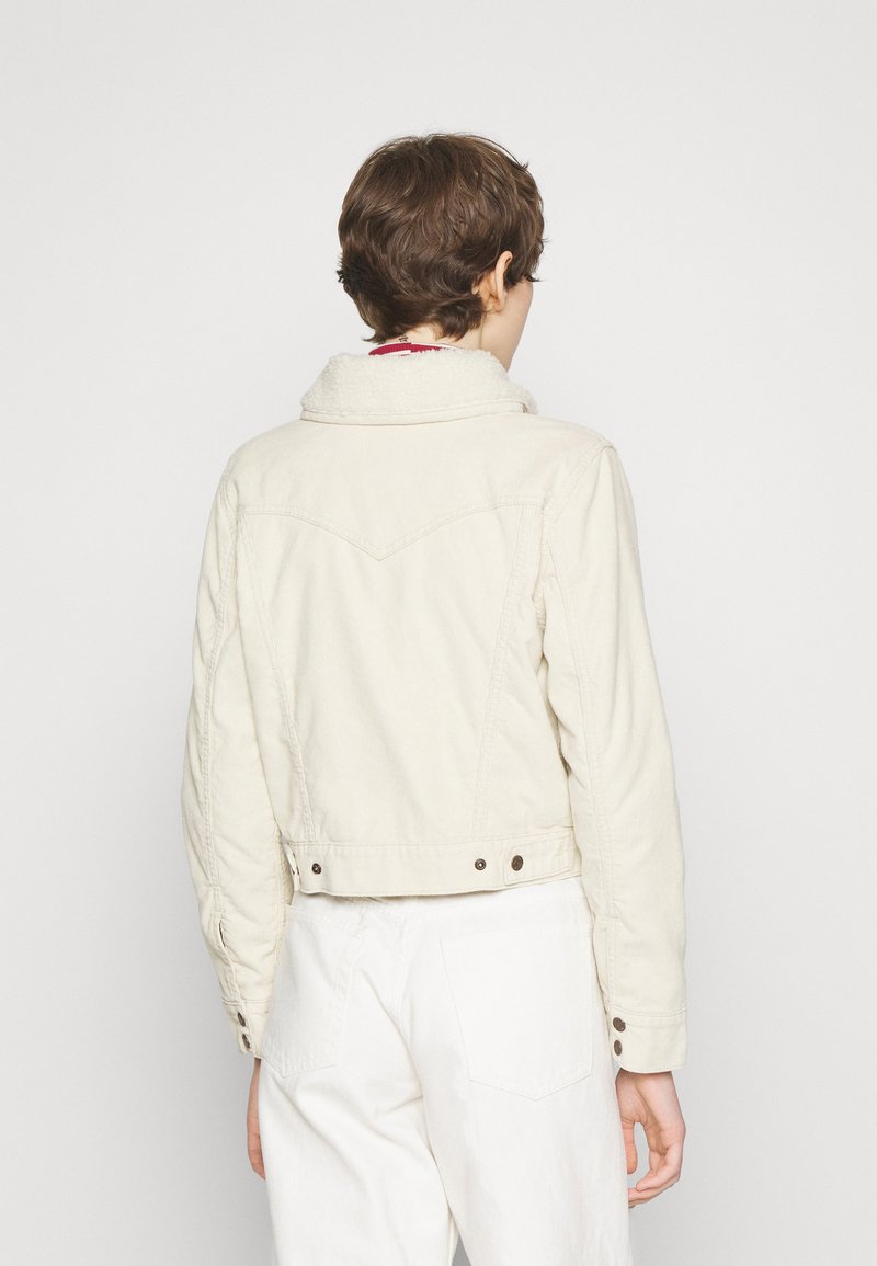 Personne avec les cheveux bruns courts portant une veste en sherpa de couleur crème et un pantalon blanc, de dos contre un fond uni.