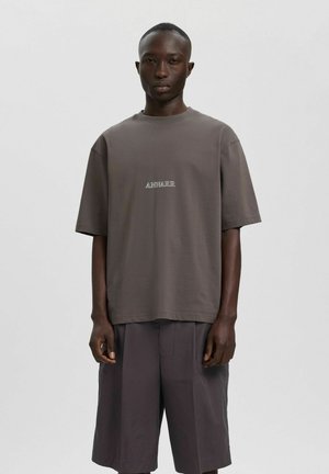 Noor mustanah mees seisab, kandes lahtist halli t-särki tekstiga "ANNARK" ja sobivaid laiu tumehalli lühikesi pükse, taustaks on lihtne taust.