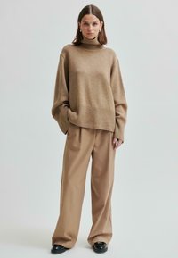 Donna che indossa un maglione beige a collo alto largo, pantaloni beige a gamba larga, scarpe nere e orecchini argentati in piedi contro uno sfondo neutro.