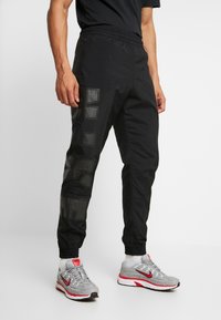 Man i svarta nylonjoggers med stora, glänsande bokstäver på benet och grå Nike-sneakers med röda detaljer mot en vit bakgrund.