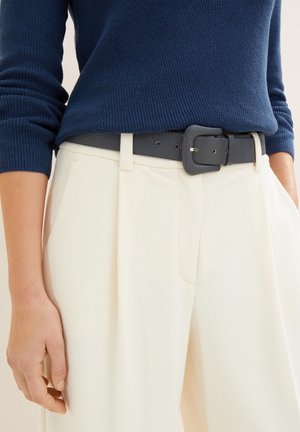 Personne portant un pantalon plissé crème taille haute avec une ceinture noire texturée et un pull côtelé à manches longues bleu marine.