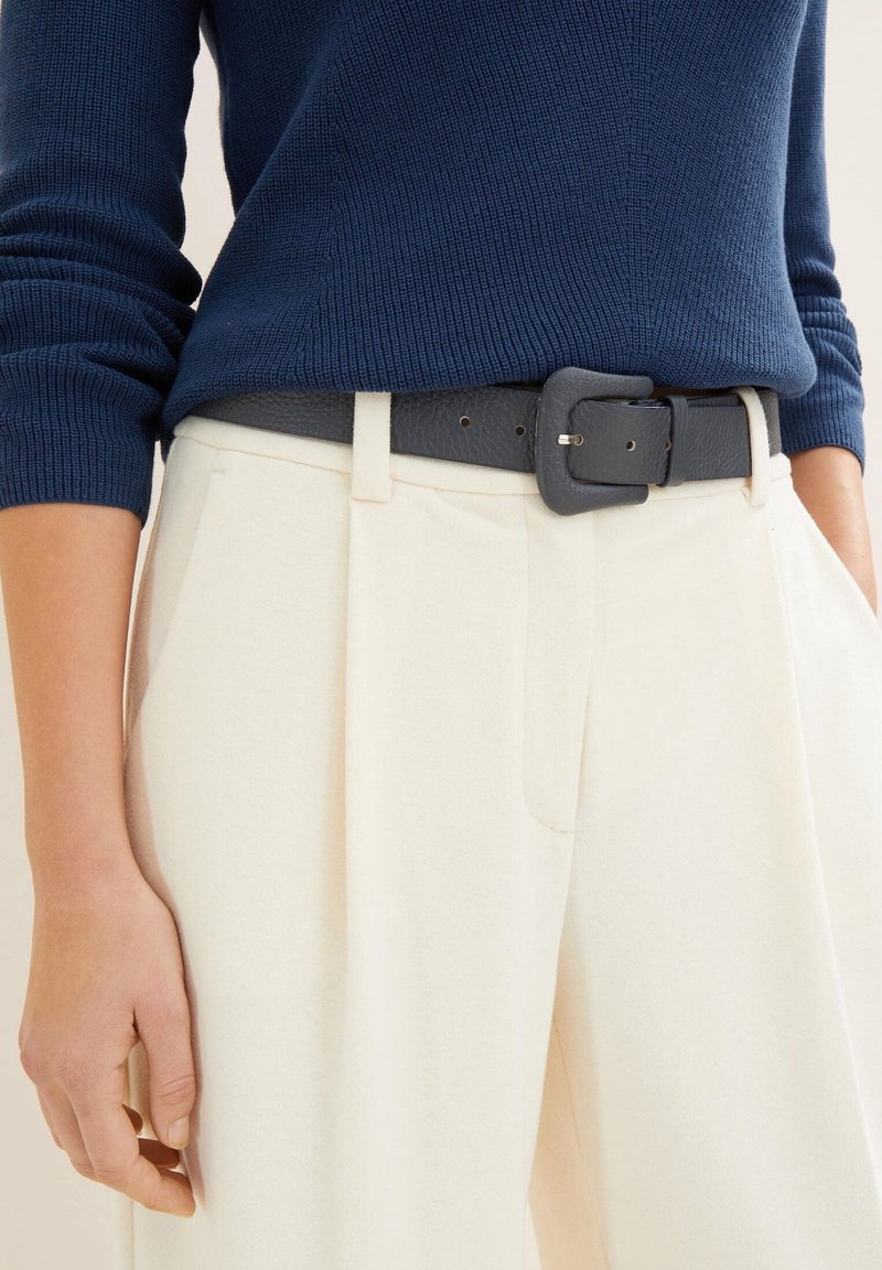 Personne portant un pantalon plissé crème taille haute avec une ceinture noire texturée et un pull côtelé à manches longues bleu marine.