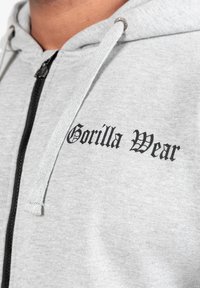 Grijze zip-up hoodie met zwarte "Gorilla Wear" tekst, gemaakt van zachte stof, met verstelbare koorden en een stevige rits.