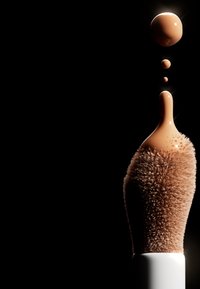 Makeupborste med mjuka, täta, bruna borst; vit handtag. Droppar av flytande foundation i en varm beige nyans sväva ovanför borsten.