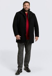 Zwarte jas met een capuchon, voorzien van een knoopsluiting aan de voorkant, twee zijzakken en een doorgestikte voering, gedragen over een rode trui en grijze broek.