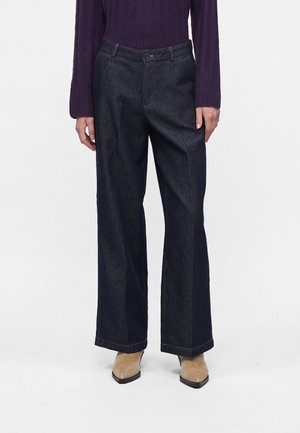 Personne portant un jean large foncé, des bottines beiges à bout pointu et un haut violet côtelé à manches longues, debout devant un fond blanc.