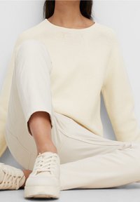 Helle cremefarbene Strickjacke mit rundem Ausschnitt, kombiniert mit beigen Slim-Fit-Hosen und passenden Sneakers. Weicher Stoff, lässige Passform.