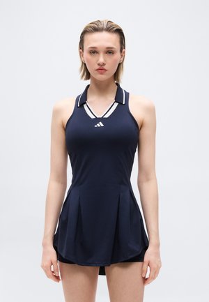 Giovane donna che indossa un vestito sportivo senza maniche blu navy con un colletto e una gonna plissettata, in piedi contro uno sfondo chiaro e uniforme.